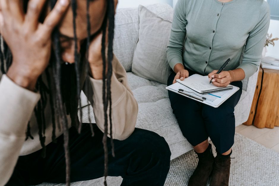 Crop concerned African American male sitting on sofa with female psychologist and holding head in hands in dismay during psychotherapy session