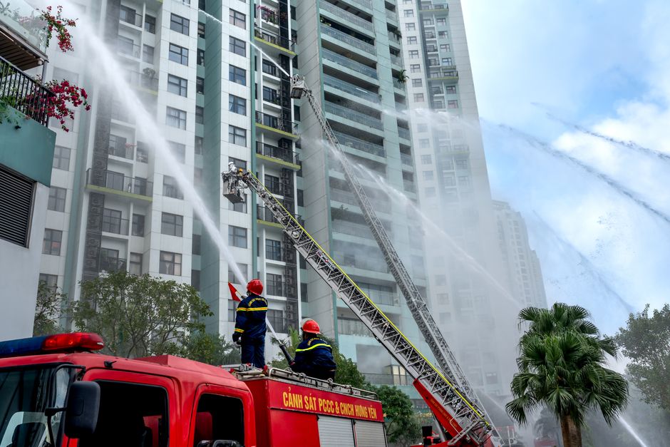 Firefighters using hoses to extinguish flames on high-rise building exterior in an urban environment.