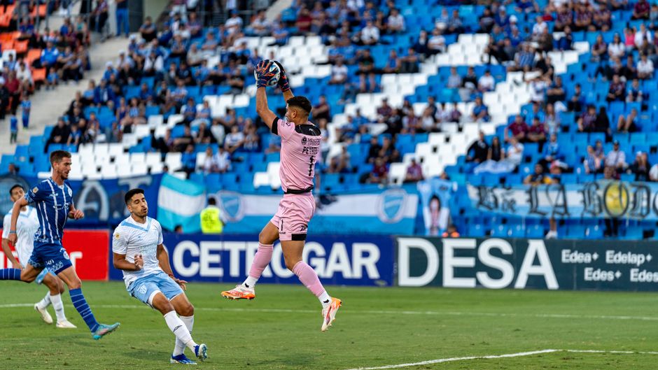 Dramatic action shot of a goalkeeper catching the ball during a competitive soccer match on a bright day.