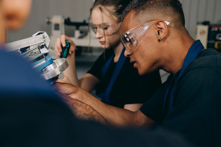 Two young adults working attentively in a workshop, wearing protective goggles.