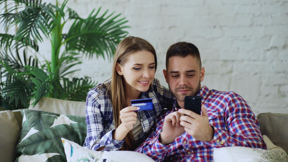 Smiling couple sitting on a sofa, shopping online with a smartphone and credit card.