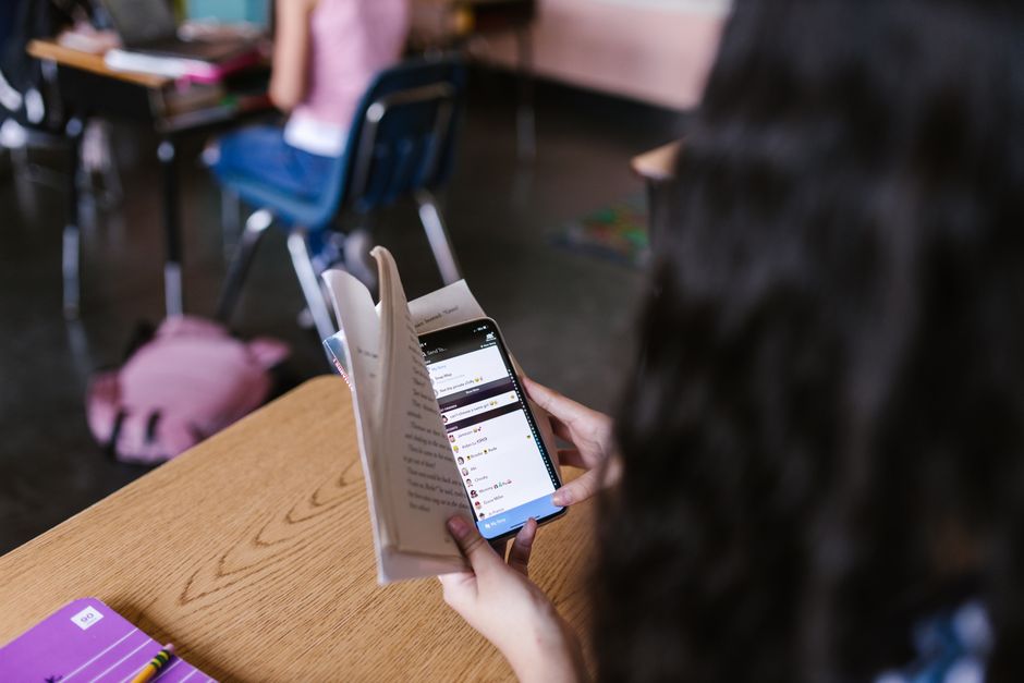 A student in a classroom multitasks by reading a book while using a smartphone, emphasizing modern educational challenges.