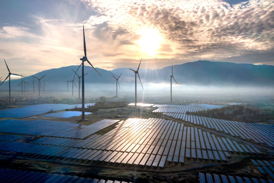 Stunning aerial shot of wind turbines and solar panels at sunrise with mountains and mist in the background.