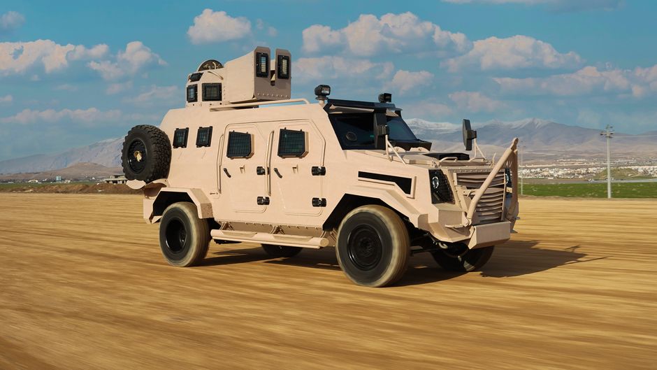 An armored military vehicle drives on a dirt road with mountain backdrop in Kurdistan.