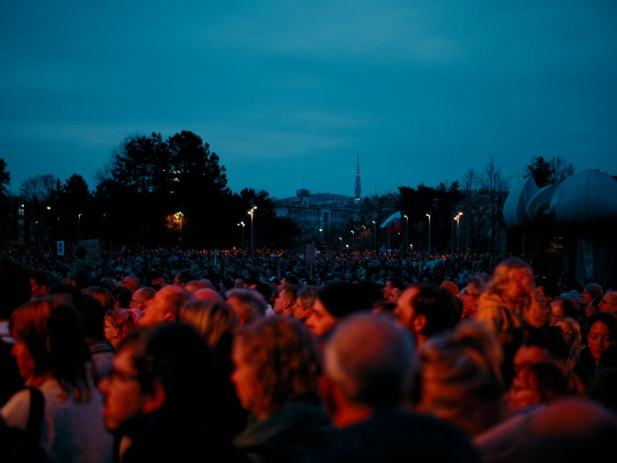 A large diverse crowd gathers outdoors at dusk for a public event, cityscape backdrop.