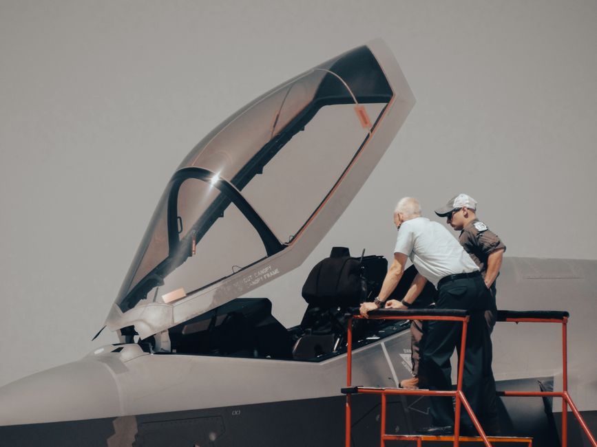 Two men inspecting military aircraft cockpit on a clear day.