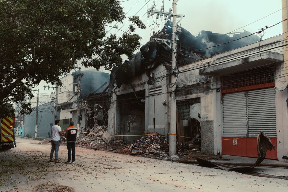 Two adults assess damage after an industrial warehouse fire. Smoke and debris visible.