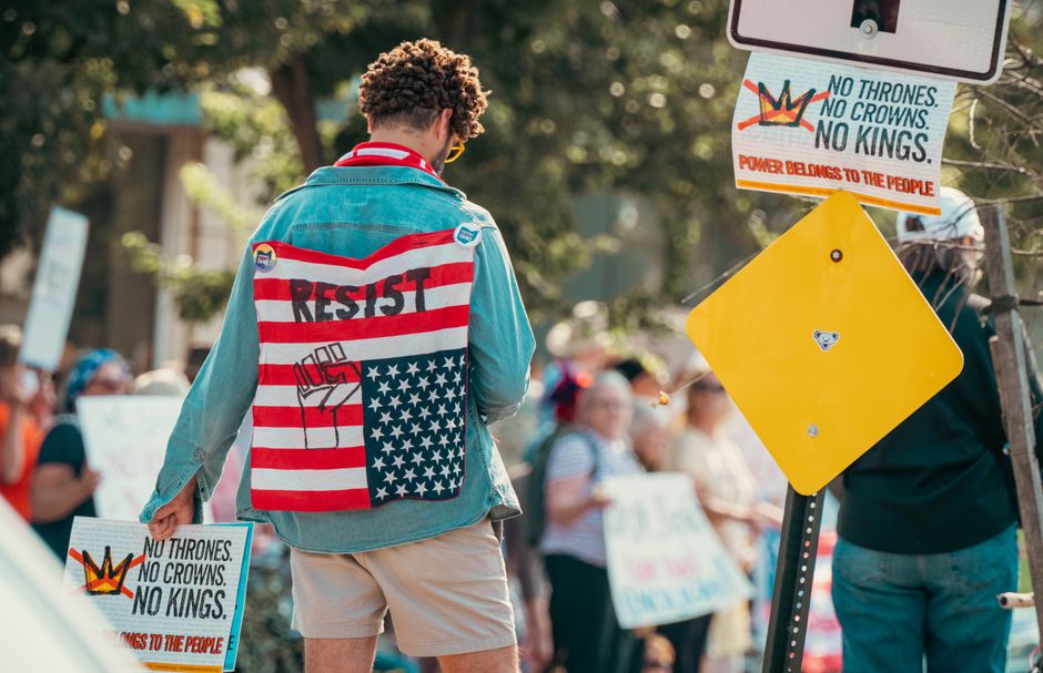 Person wearing flag-themed denim jacket at a protest holding signs promoting resistance and anti-establishment views.