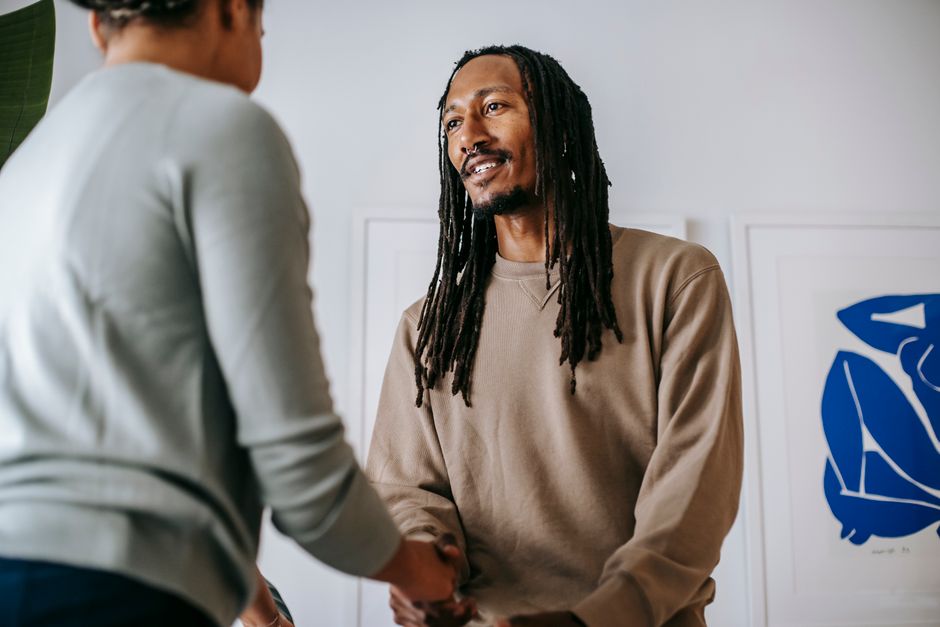 From below of cheerful bearded African American male patient with dreadlocks shaking hand of unrecognizable female therapist while standing in office during appointment