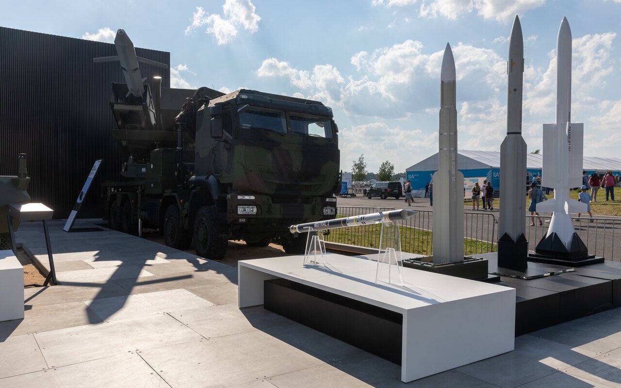 Trois missiles blancs dressés sur un socle d'exposition, véhicule lanceur sombre en arrière-plan