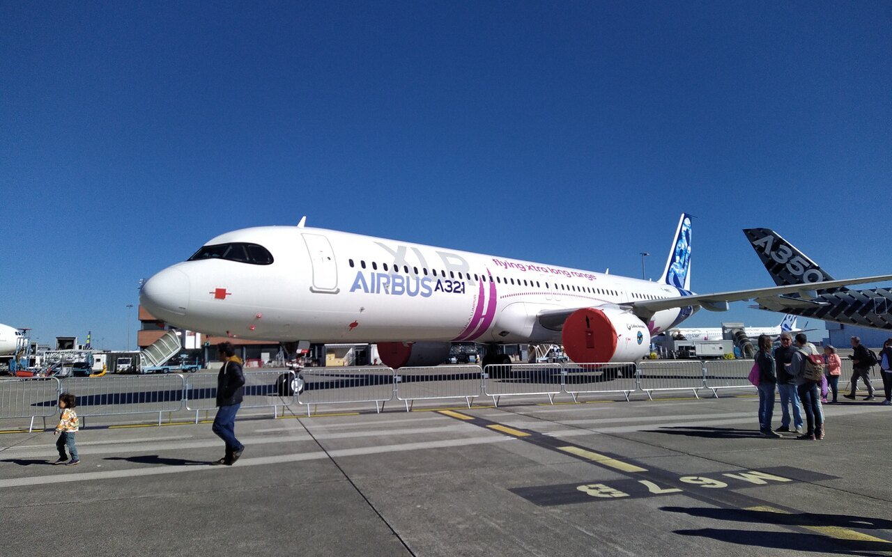 Un avion de ligne blanc aux marques du constructeur, stationné sur une aire devant des visiteurs