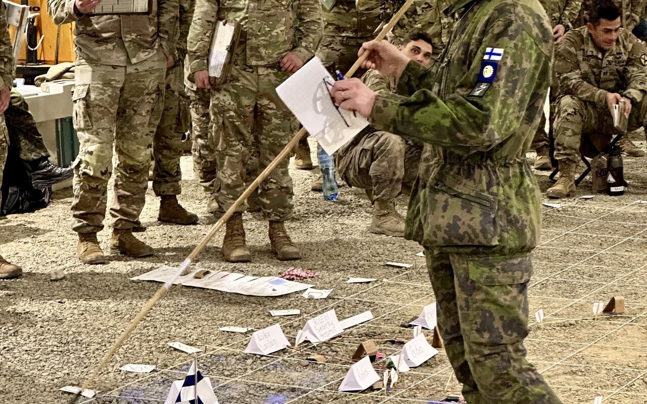 Des militaires rassemblés autour d'une maquette au sol faite de papiers, un officier briefant debout