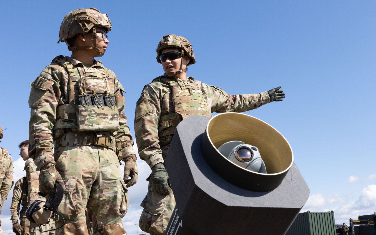 Deux militaires debout à côté d'un tube de lancement hexagonal dont on voit la gueule, au bord d'un terrain côtier