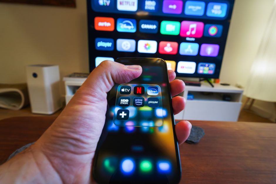 Main tenant un téléphone affichant des applications de streaming devant un écran de télévision.