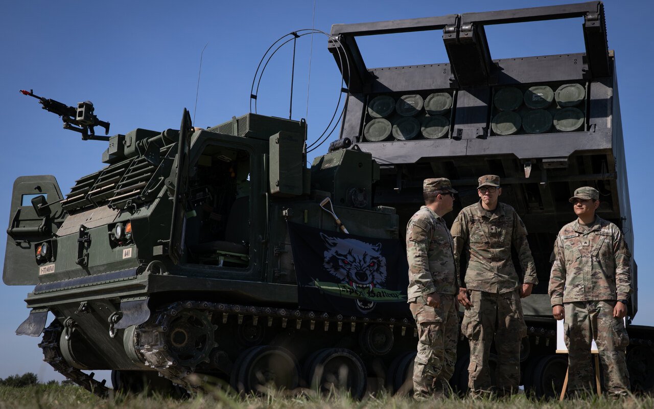 Trois militaires en tenue de combat discutent au pied d'un lance-roquettes chenillé, conteneur de roquettes visible