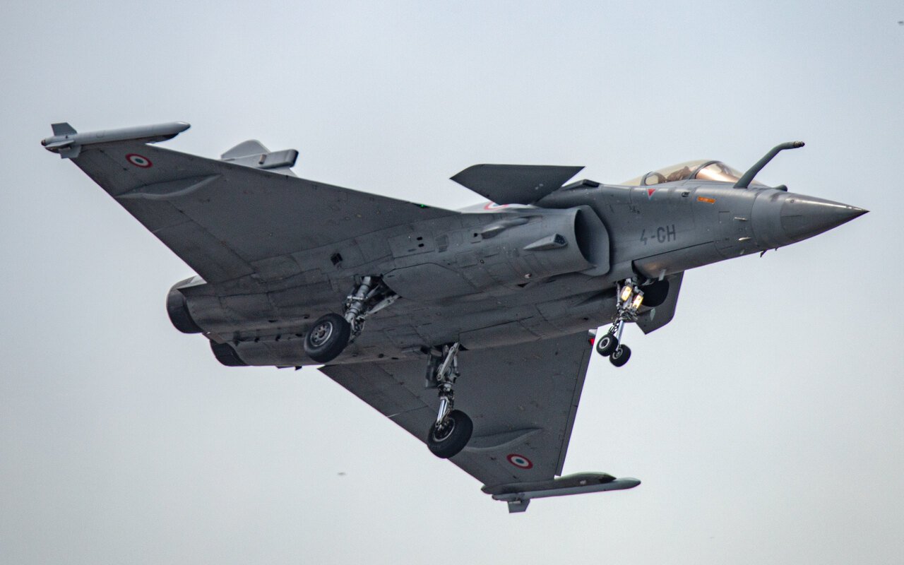 Un avion de combat gris vu de dessous en vol, train sorti, sous un ciel laiteux