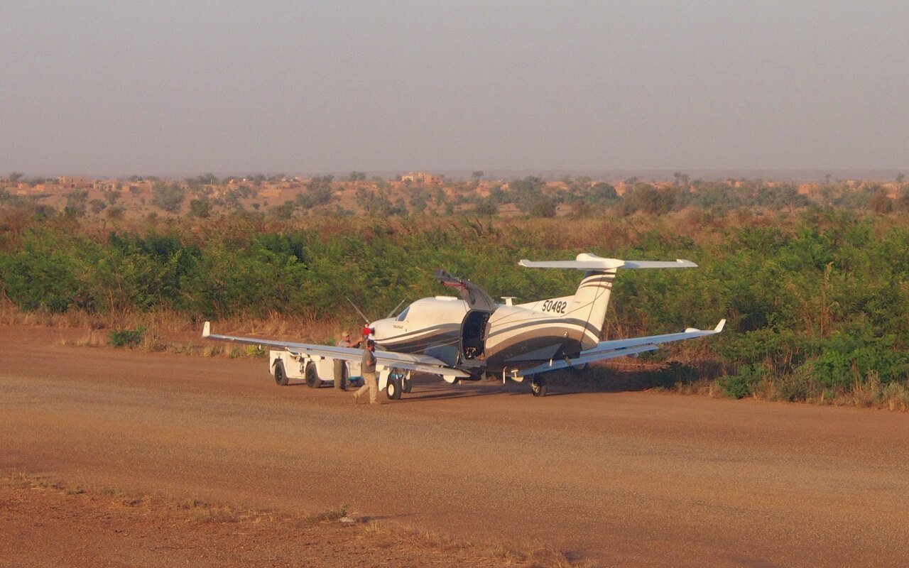 Un avion léger beige posé sur une piste de latérite, végétation sèche et ciel chargé de poussière