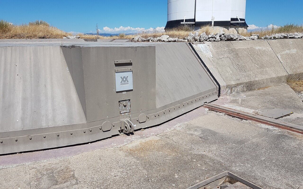 Une épaisse dalle de béton basculée sur ses rails au-dessus d'un silo désaffecté, végétation sèche alentour